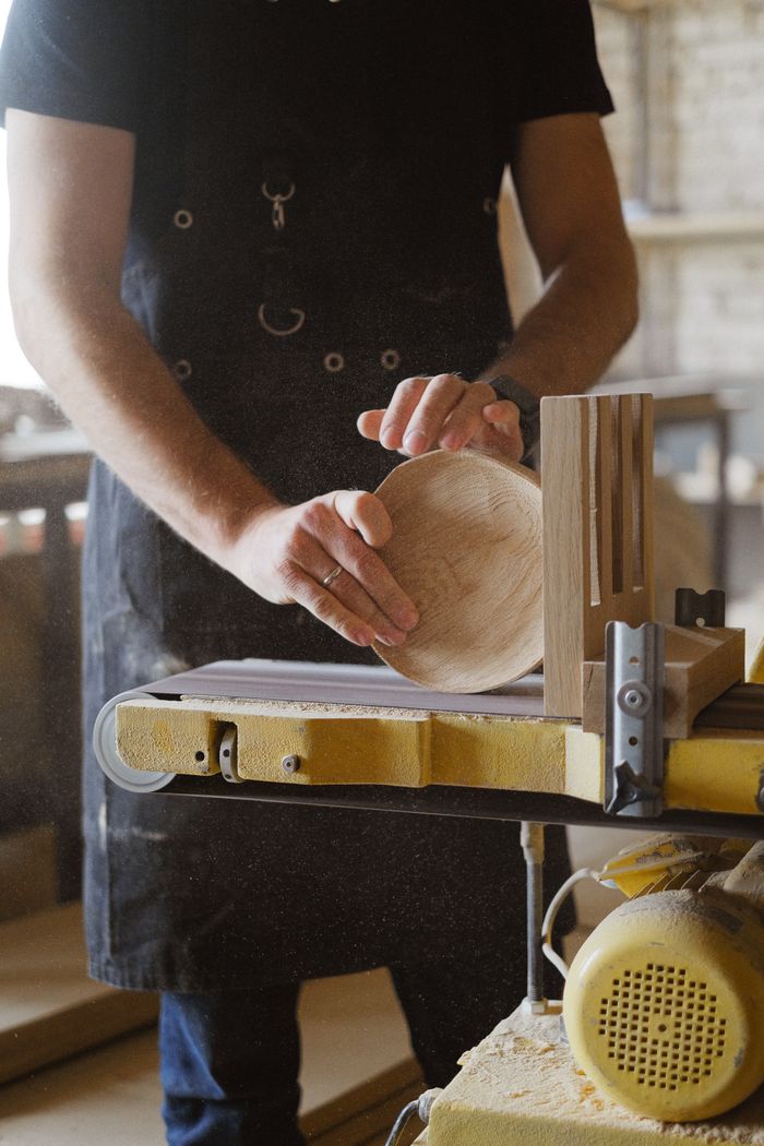 Unrecognizable male master in black apron using special instrument to grind round piece of wood while working in professional carpentry