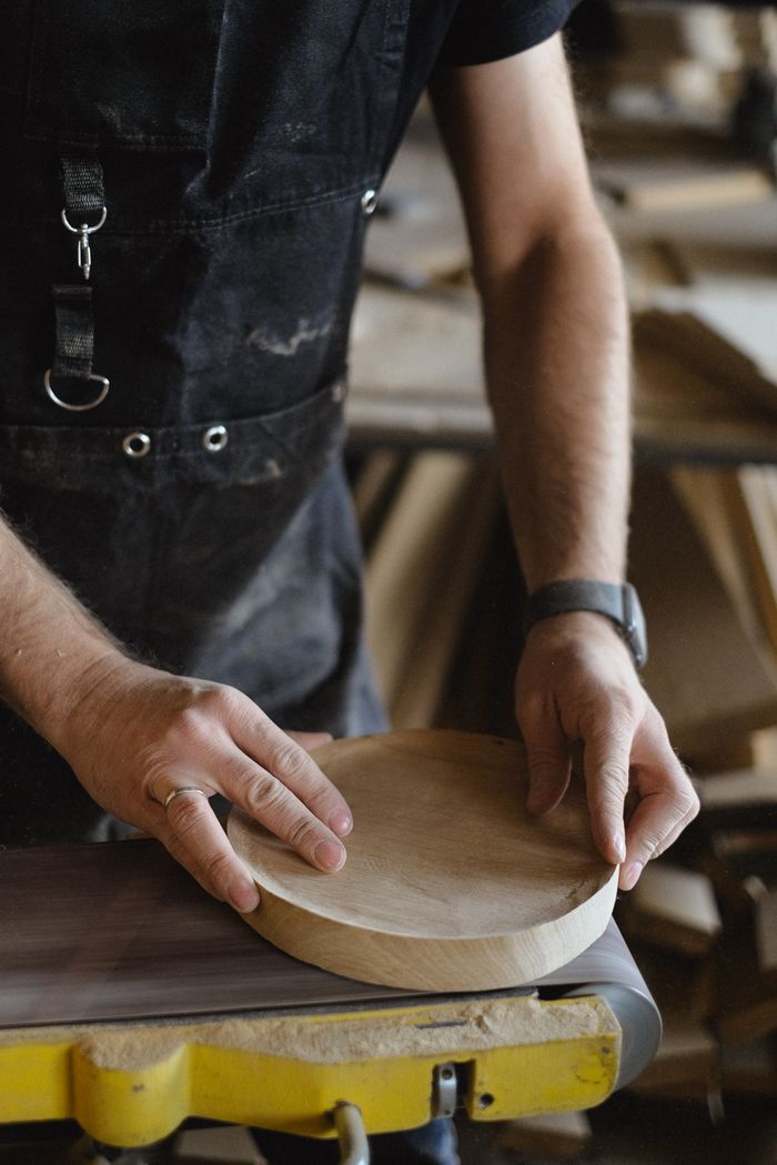 Unrecognizable carpenter using belt and disc sander to shape round piece of wood while working in professional joinery on blurred background