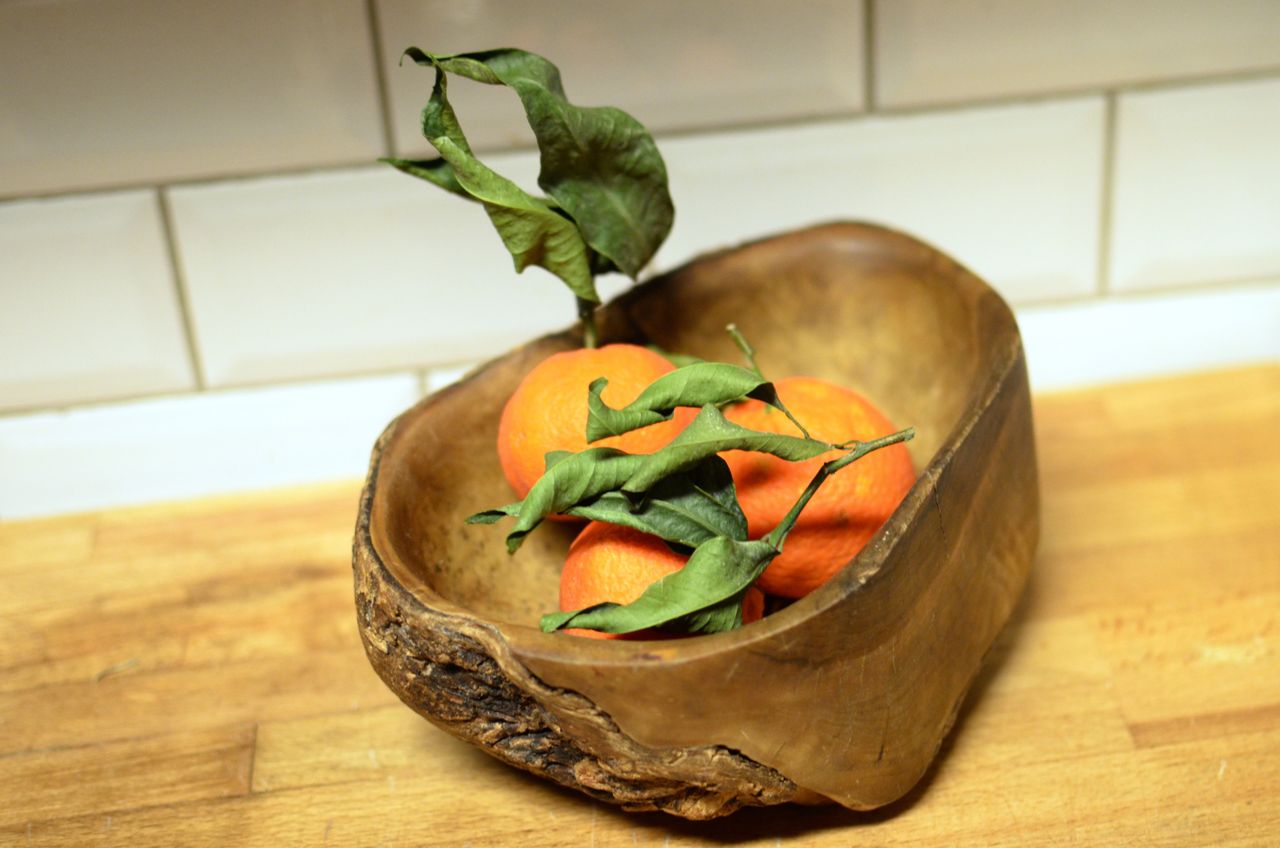 Fresh oranges with green leaves in a wooden bowl on a wooden surface, perfect for rustic kitchen themes.